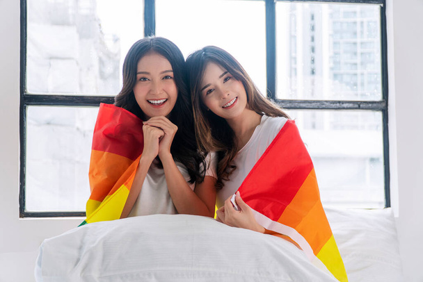 Romantic asian couple woman hugging together with rainbow flag sitting on bed beside window. Cheerful lover lesbian casual girl with lgbt flag hug closeness at home. Lgbt couple relationship concept. - Foto, Imagem