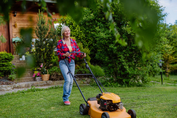 庭の芝刈り機で草を刈る年配の女性、庭の仕事の概念. - 写真・画像
