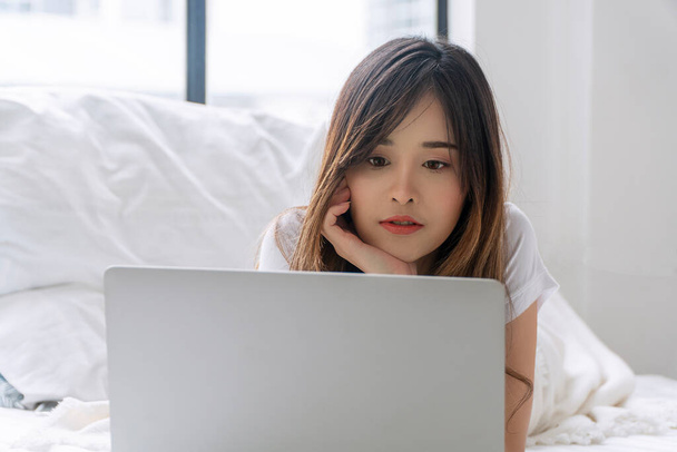 Freelance asian woman looking laptop lying down on white bed working at home. Teenager young girl using notebook learning online or shopping online or social media entertainment in morning. Technology - Valokuva, kuva