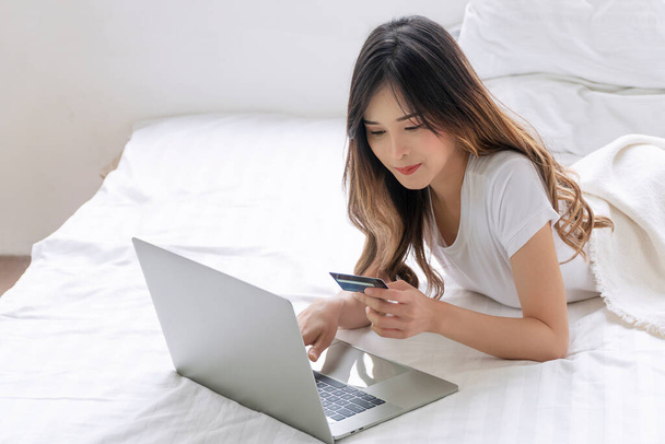 Cheerful asian woman holding credit card or debit mockup electronic payment shopping online with laptop lying on white bed at home. Happy young girl using mockup card pay banking e-commerce.Technology - Fotó, kép