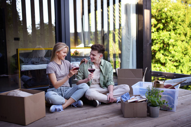 Ein fröhliches junges Paar zieht in sein neues Häuschen im Wald, sitzt auf dem Fußboden auf der Terrasse und trinkt Wein. Konzept des bewegten und nachhaltigen Lebens. - Foto, Bild