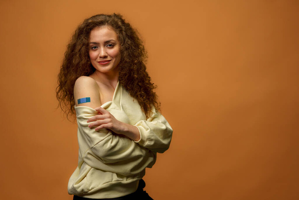 A portrait of a female smiling after getting a vaccine. Woman holding down her shirt sleeve and showing her arm with bandage after receiving vaccination isolated on orange background. - Foto, immagini