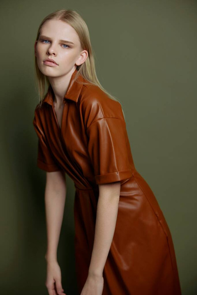 Stylish young blonde model with blue eyes and evening makeup posing in trendy leather fashion brown palette clothes leaning forward looks at camera. Studio portrait isolated on olive green background - Photo, Image