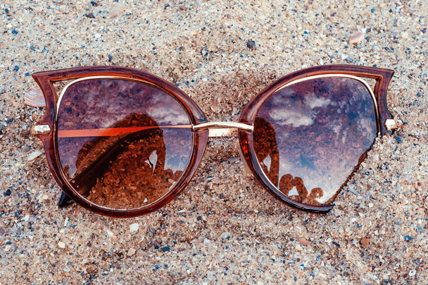 This photo depicts image of Top view of sunglasses on the beach near the blue sea with sand. - Zdjęcie, obraz