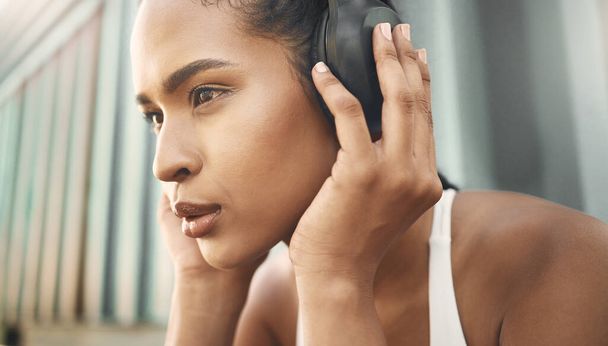 Closeup of one fit young hispanic woman listening to music with headphones while exercising in an urban setting outdoors. Face of focused and motivated female athlete ready for training workout or run - 写真・画像