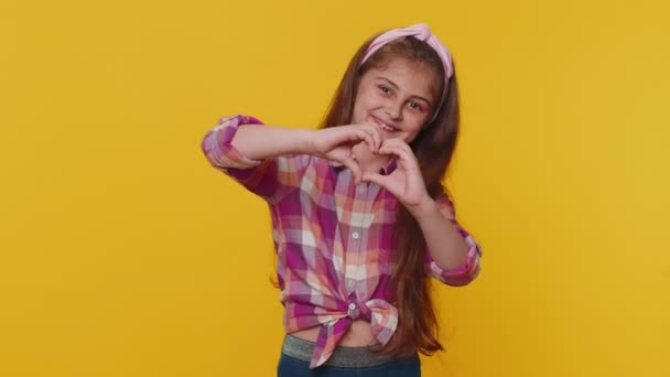 I love you. Smiling young preteen child girl kid makes heart gesture demonstrates love sign expresses good feelings and sympathy. Little toddler children isolated alone on studio yellow background - Materiał filmowy, wideo