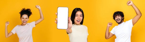 Foto panorámica de tres personas multirraciales sobre fondo naranja aislado. Chica asiática muestra teléfono inteligente con pantalla de maqueta blanca en blanco, chico indio y chica afroamericana celebrando el éxito, sonrisa - Foto, imagen