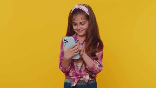 Młody nastolatek dziecko dziewczyna używać mobilny smartfon wpisując przeglądanie powiedzieć Wow tak okazało się wielkie zwycięstwo dobre wieści świętować robi gest zwycięzca. Małe maluszki na żółtym tle studio - Materiał filmowy, wideo