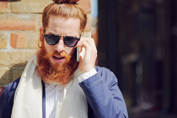 One trendy young fashionable man with man bun wearing sunglasses and talking on a cellphone while standing outside enjoying the sun in the city. Caucasian man with a full beard against a brick wall. - Foto, imagen