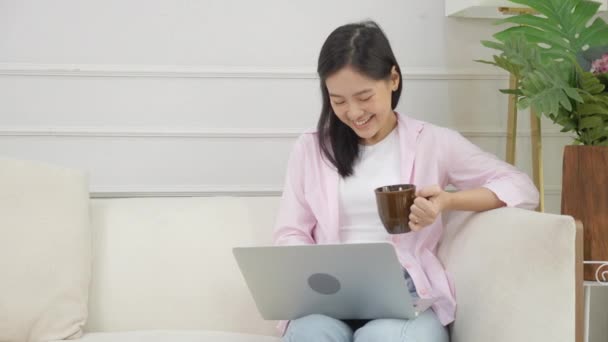 Joven mujer de negocios asiática trabajar desde casa con ordenador portátil y beber café en el sofá en la sala de estar, chica independiente utilizando portátil sentado en el sofá con comodidad y relax, el concepto de estilos de vida. - Metraje, vídeo
