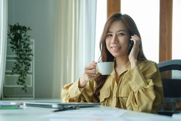 Millennial Ασίας επιχειρηματίας κρατώντας φλιτζάνι καφέ και μιλώντας στο κινητό τηλέφωνο. - Φωτογραφία, εικόνα
