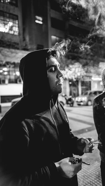 Young latino / caucasian guy smoking a joint (marijuana cigarette, pot cigarette) and a tobacco cigarette, blowing smoke in the street in the night - Photo, Image