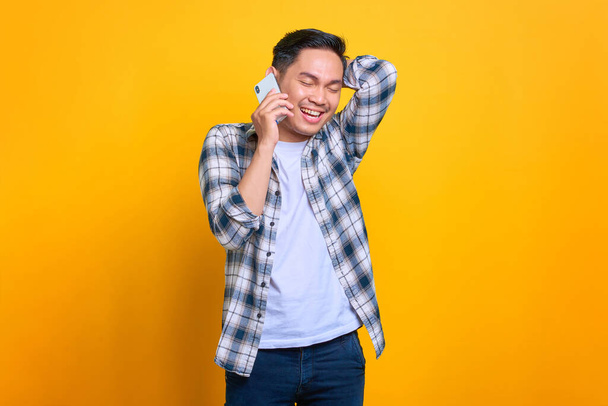 Cheerful young Asian man in plaid shirt talking on mobile phone isolated on yellow background - Fotoğraf, Görsel