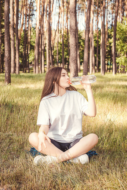 Deze foto toont beeld van Een mooi jong meisje drinkt water uit een fles na training voor verfrissing in de tuin, ontspant en traint. - Foto, afbeelding
