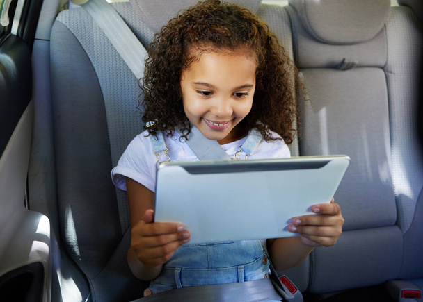 Vágott kép egy imádnivaló kislányról, aki a tabletjét használja, miközben egy autó hátsó ülésén ül.. - Fotó, kép