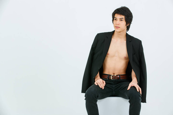 Portrait isolated studio shot Asian young handsome confident slim thin shirtless healthy athletic teen fashion male model in suit blazer jeans and sneakers sitting smiling posing on white background. - 写真・画像