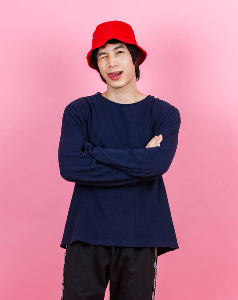 Portret studio schot aziatische jong stedelijk tiener mode man model in straat stijl outfit met lange mouw shirt rood emmer hoed staande gekruiste armen glimlachende blik op camera op roze achtergrond - Foto, afbeelding