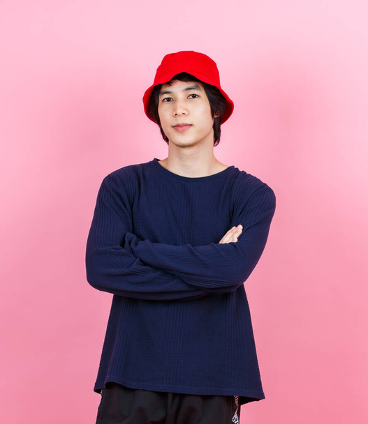 Portrait studio shot Asian young urban teen fashion male model in street style outfit long sleeve shirt red bucket hat standing crossed arms smiling look at camera on pink background - Photo, Image