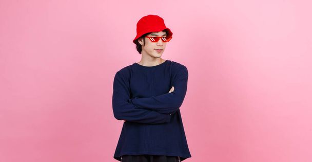 Portrét studio shot asijské mladý městský dospívající móda muž model v ulici styl outfit dlouhý rukáv košile červená vědro klobouk legrační brýle stojící zkřížené paže úsměv pohled na kameru na růžovém pozadí. - Fotografie, Obrázek