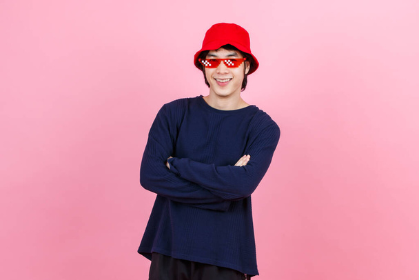 Portrait-Studio erschossen asiatische junge urbane Teenager-Mode männliches Modell in Street-Style-Outfit langärmeliges Hemd roter Eimer Hut lustige Brille stehend verschränkte Arme lächelnd in die Kamera auf rosa Hintergrund. - Foto, Bild