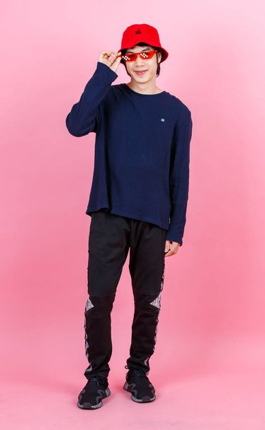 Portrait studio shot Asian young urban teen fashion male model in street style outfit long sleeve shirt red bucket hat funny glasses standing crossed arms smiling look at camera on pink background. - Foto, Imagem
