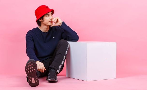 Portrait studio shot of Asian young urban teen fashion male model in street style outfit long sleeve shirt sport pants sneaker red bucket hat sitting relaxing leaning at white box on pink background. - Photo, Image