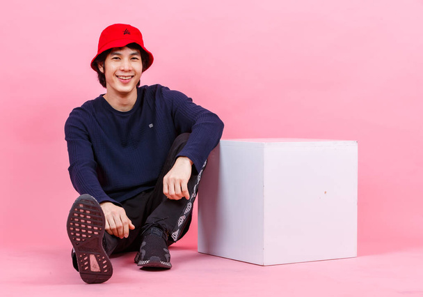 Portrait studio shot of Asian young urban teen fashion male model in street style outfit long sleeve shirt sport pants sneaker red bucket hat sitting relaxing leaning at white box on pink background. - 写真・画像