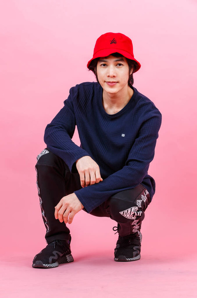 Portrait studio shot of Asian young urban teen fashion male model in street style outfit long sleeve shirt sport pants sneaker red bucket hat sitting relaxing leaning on pink background. - Photo, image