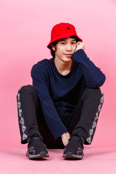 Πορτρέτο στούντιο πλάνο της Ασίας νεαρός urban έφηβος μόδας ανδρικό μοντέλο σε στυλ δρόμου στολή μακρύ μανίκι πουκάμισο σπορ παντελόνι sneaker κόκκινο καπέλο κουβά κάθεται χαλαρωτικό κλίνει σε ροζ φόντο. - Φωτογραφία, εικόνα
