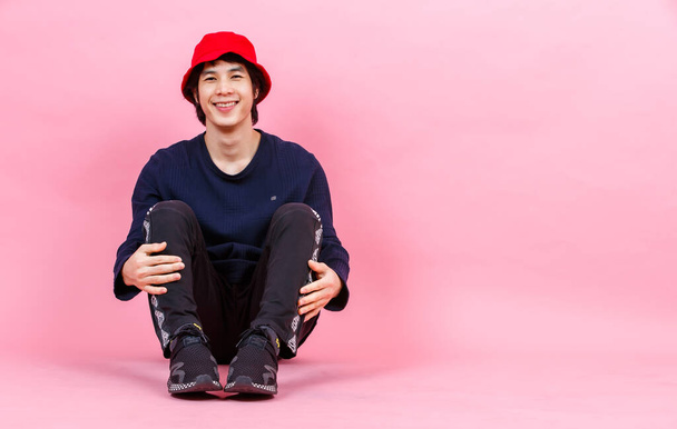 Πορτρέτο στούντιο πλάνο της Ασίας νεαρός urban έφηβος μόδας ανδρικό μοντέλο σε στυλ δρόμου στολή μακρύ μανίκι πουκάμισο σπορ παντελόνι sneaker κόκκινο καπέλο κουβά κάθεται χαλαρωτικό κλίνει σε ροζ φόντο. - Φωτογραφία, εικόνα