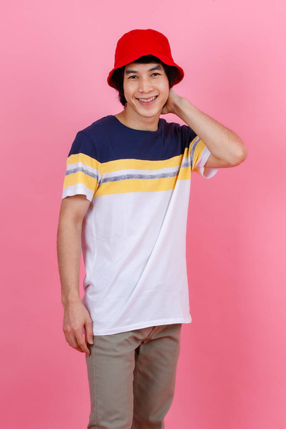 Portrait studio shot of Asian young handsome urban teenager fashion male model in street style outfit t shirt sport beige pants and red bucket hat standing posing look at camera on pink background. - Photo, image