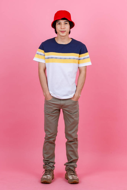 Portrait studio shot of Asian young handsome urban teenager fashion male model in street style outfit t shirt sport beige pants and red bucket hat standing posing look at camera on pink background. - Foto, Bild