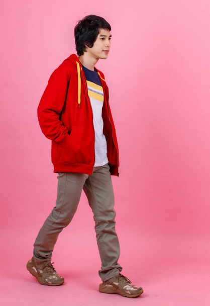 Portret pełne ciało studio strzał Azji młody miejski nastolatek moda męski model w stylu ulicy strój czerwony kurtka stoi pozuje trzymając się za ręce w spodniach kieszenie patrzeć na aparat fotograficzny na różowym tle. - Zdjęcie, obraz