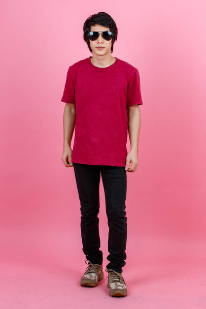 Portrait studio shot of Asian young urban teenager fashion male model in casual street style outfit t shirt and black jeans with aviator sunglasses standing posing look at camera on pink background. - Photo, image