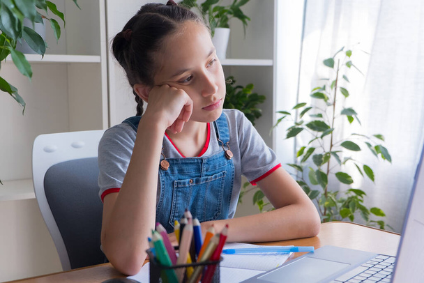 Trieste brunette 11-13 jaar oud, gekleed in een grijs T-shirt, zit thuis aan tafel met een computer, doet huiswerk, studies, communiceert op het internet. - Foto, afbeelding