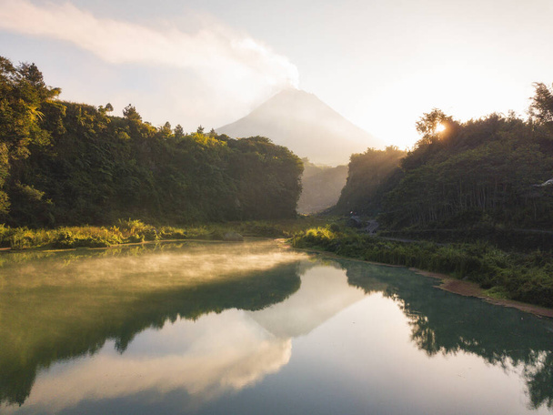 日の出を背景に火山で朝の煙を放出し、緑に見える水面を持つ湖。その湖は木に囲まれている。インドネシアのメラピ火山という火山. - 写真・画像