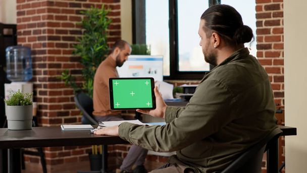 Pracownik firmy patrzy na tablet z poziomym zielonym ekranem na wyświetlaczu, pracuje nad biznesem z tłem klucza chroma i odizolowanym szablonem copyspace. Blank mockup ekranu w biurze. - Zdjęcie, obraz