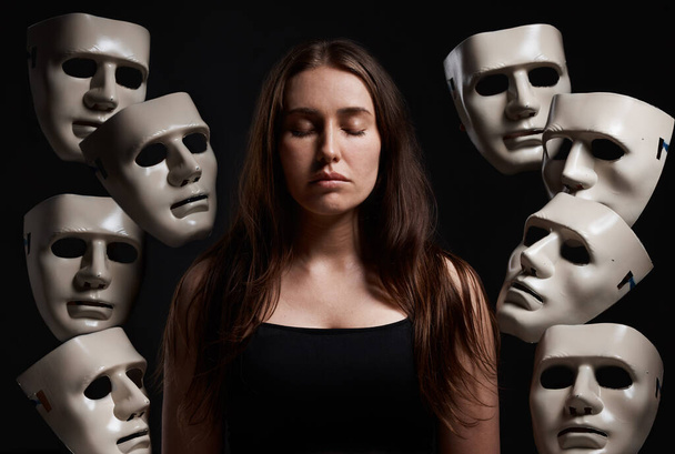 Studio shot of a woman standing with her eyes closed while surrounded by masks. - Foto, afbeelding