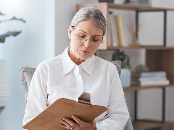 Shot of a mature psychologist sitting in her practice and writing on a clipboard. - Foto, afbeelding