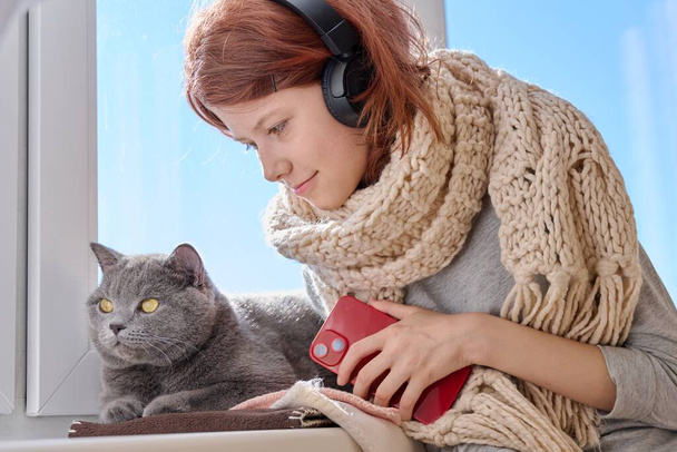 Preteen girl in knitted woolen scarf with cute gray cat near the winter window. A child in headphones with phone along with pet on winter sunny day - Foto, Bild