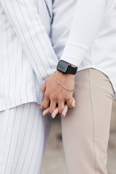 a girl and a boy sit close to each other holding hands. close-up photo of clasped hands. the guy has a modern watch and a bracelet on his hand - Foto, afbeelding