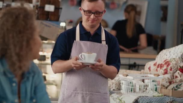 Caucasian male waiter with down syndrome serving a cup of coffee in the cafe to the client at the table. Shot with RED helium camera in 8K.  - Materiał filmowy, wideo