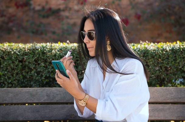 une belle jeune femme sud-américaine en vacances en Europe, est assise sur un banc de parc peignant ses lèvres tout en utilisant son téléphone portable comme miroir. - Photo, image