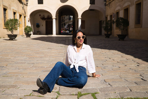 young, beautiful, brunette south american woman is sitting on the brick floor in a typical mediterranean city sunbathing. The woman poses for the photo. Holiday and travel concept - Фото, зображення