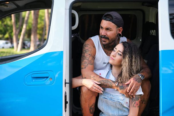 Νεαρό ζευγάρι με τατουάζ αγκαλιάζει και κοιτάζει μια πλευρά, κάθεται στο πλάι του φορτηγού. - Φωτογραφία, εικόνα