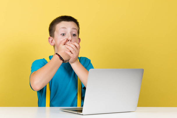 Pokutujący chłopiec trzyma ręce na ustach. Zaskoczony nastolatek przed laptopem. - Zdjęcie, obraz