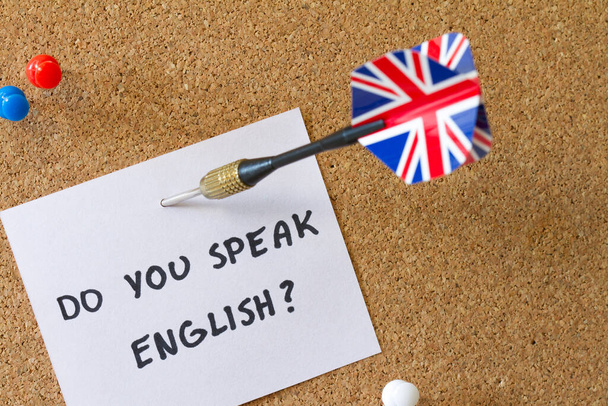 Aiming with dart with Uk flag at text "do you speak english" on cork board, english language learning concept - Foto, Imagen