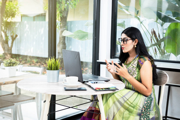 Indian woman with eyeglass using a laptop with a notebook and a mobile phone with a cup of coffee on the table in the coffee shop - Photo, image