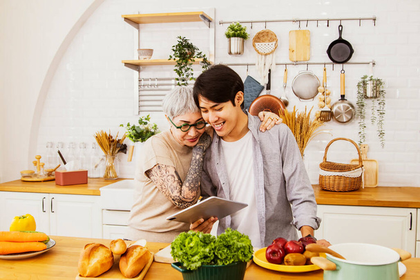 Couple between elderly woman with arm tattoos and handsome childhood boy who bond and love each other look happily looking after the laptop searching for the cooking menu in the kitchen with joy. - Φωτογραφία, εικόνα