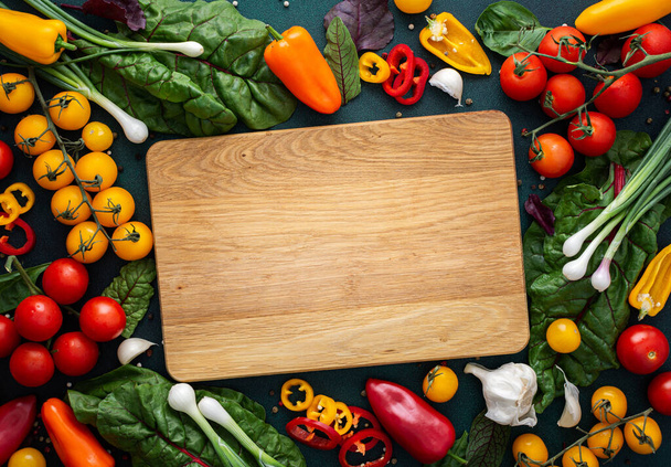 Empty cutting board in the middle of fresh organic vegetables on a green background, food background, farm products, colorful peppers and tomatoes, chard leaves and onions - 写真・画像